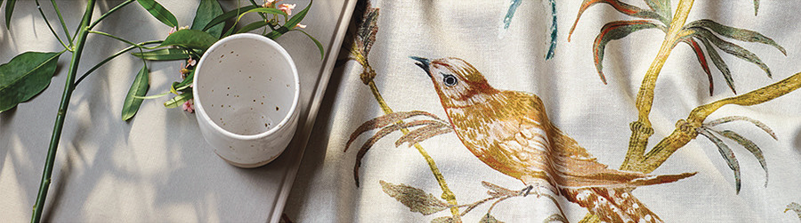 Bird print linen fabric draped on a table with foliage and ceramics – soft botanical bird fabric for curtains and upholstery